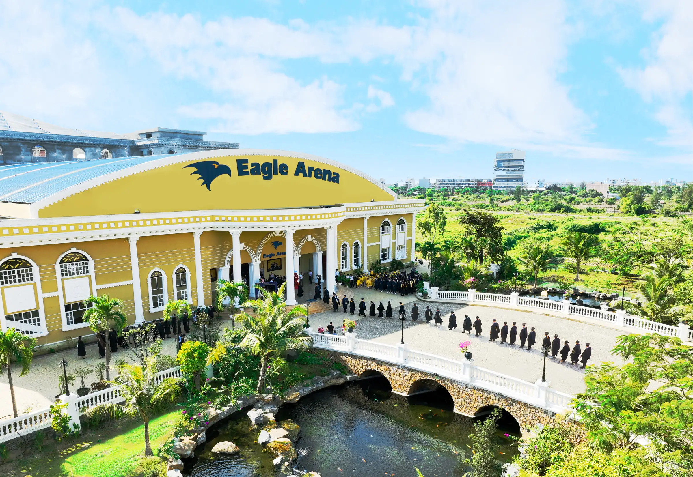 Graduates in gowns cross the stone bridge toward Eagle Arena on the AUV campus